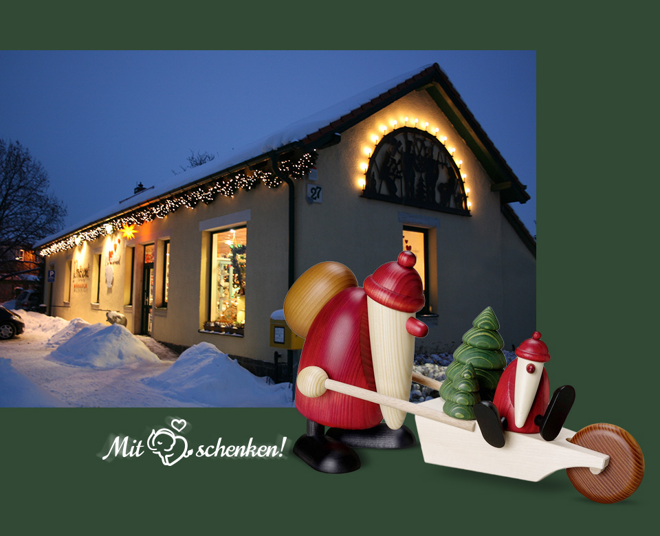 Ladengeschäft von Präsent Münnich in Ottendorf-Okrilla bei Nacht und eine traditionelle Holzkunst-Figur von Köhlers ein Weihnachtsmann mit kleinerem Wichtel und Tannenbäumen auf einer Schubkarre mit dem Text Mit Herz schenken